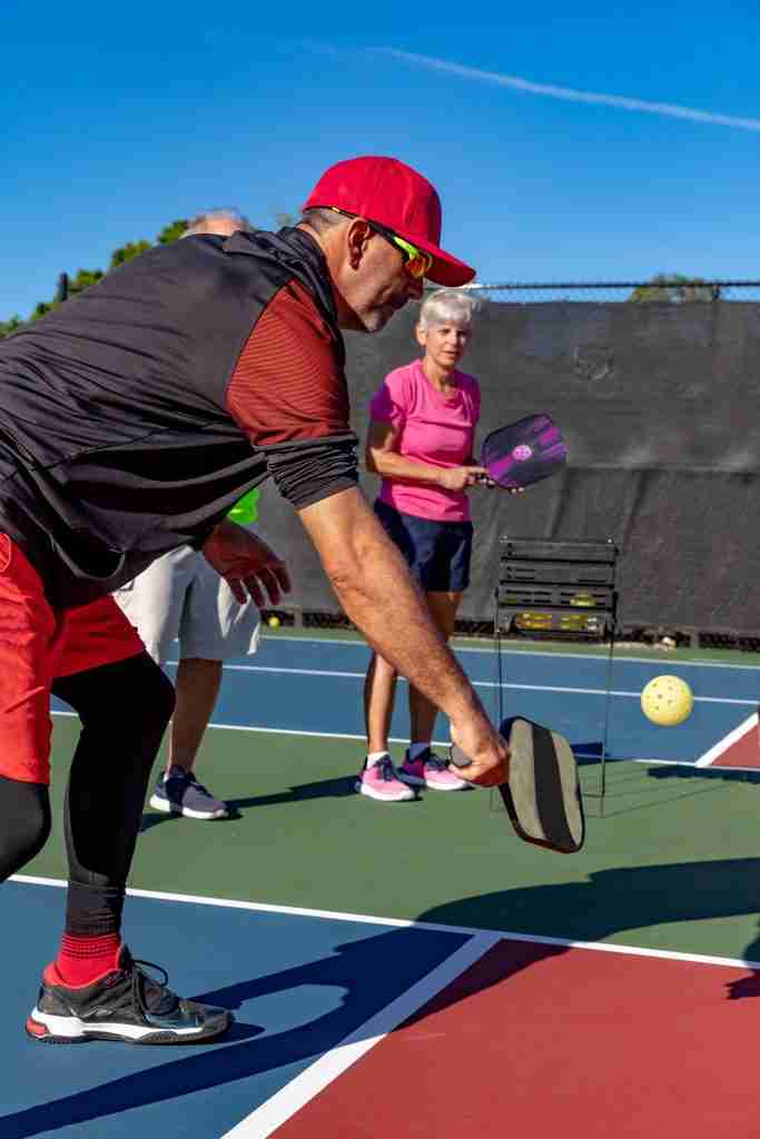 Male pickleball player in red cap demonstrating a shot technique on an outdoor court, with two players observing in the background, showcasing tips and tricks for gameplay.