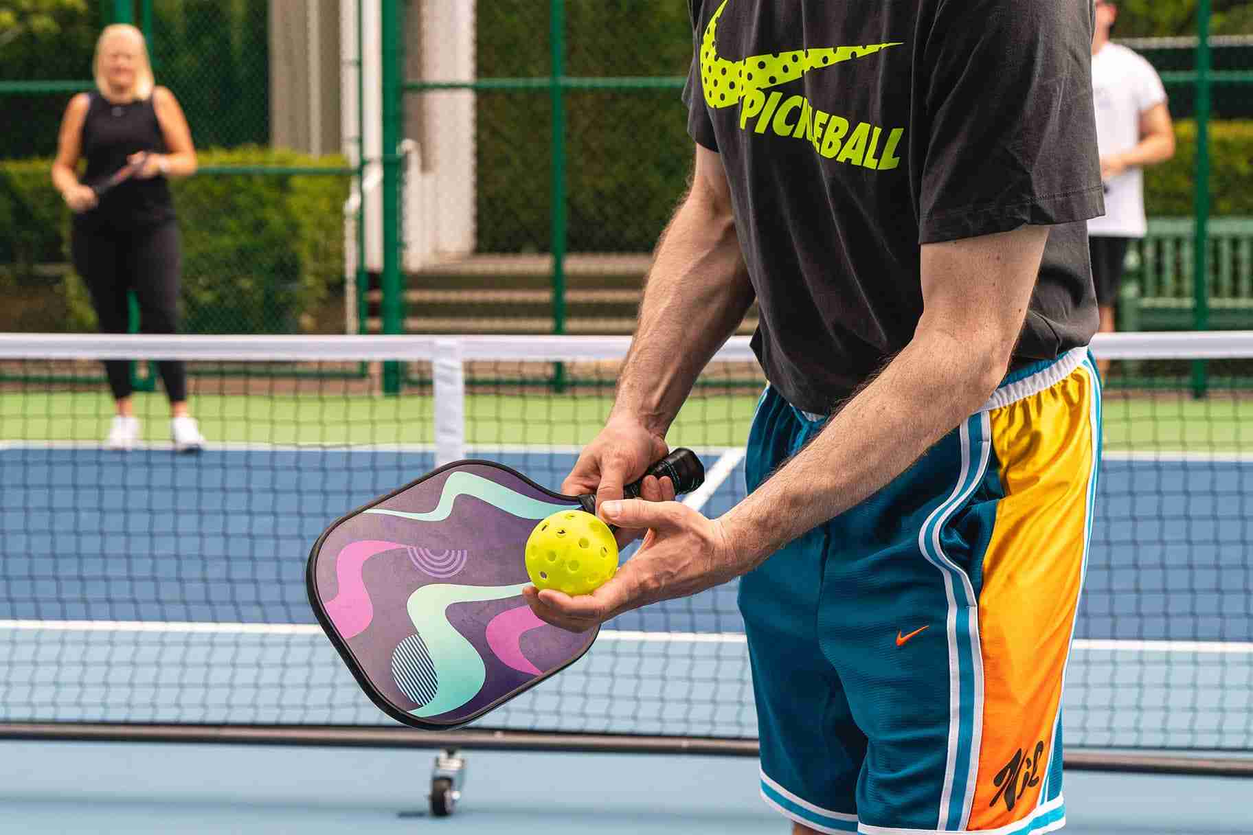 Player serving a yellow pickleball with a colorful paddle, opponent ready across the net.