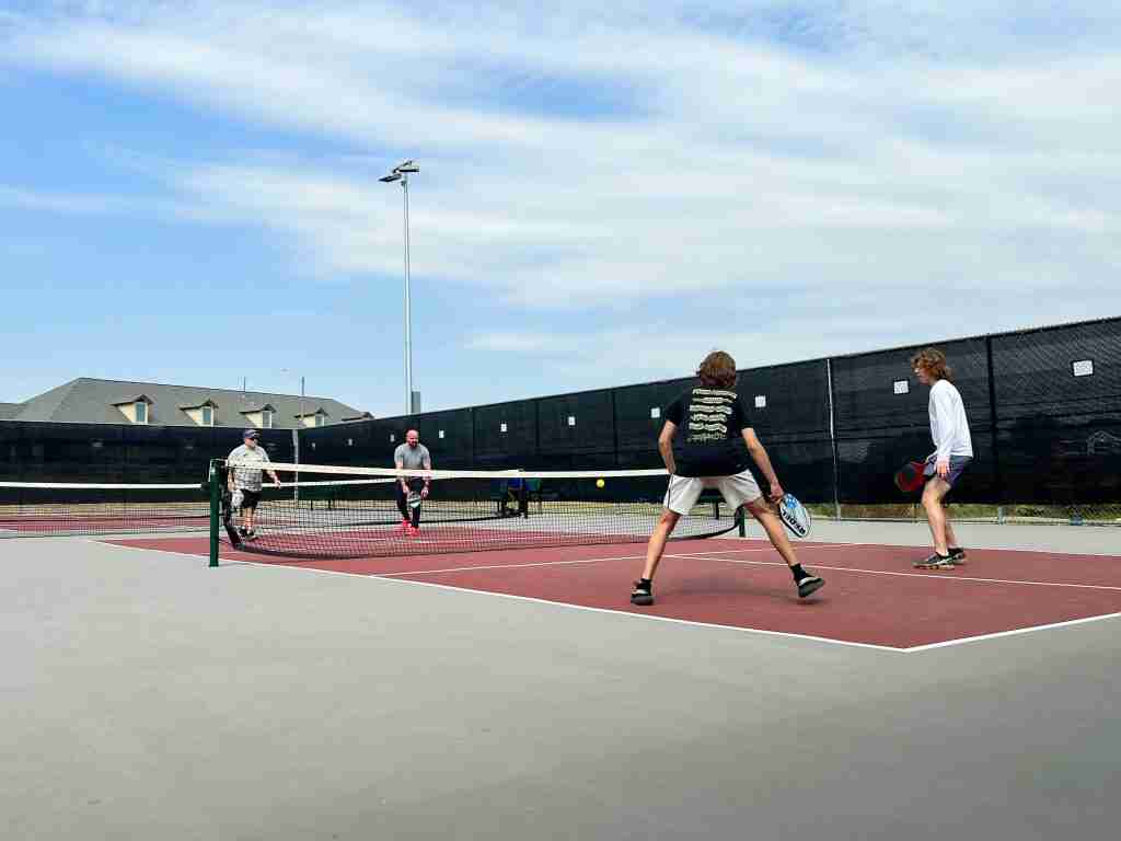 Four players engaged in a pickleball game on an outdoor court, demonstrating the double bounce rule. Two teams of two are positioned on each side of the net, actively playing. The court is marked for pickleball with players in motion, emphasizing the rule where the ball must bounce once on each side before volleys can be played.