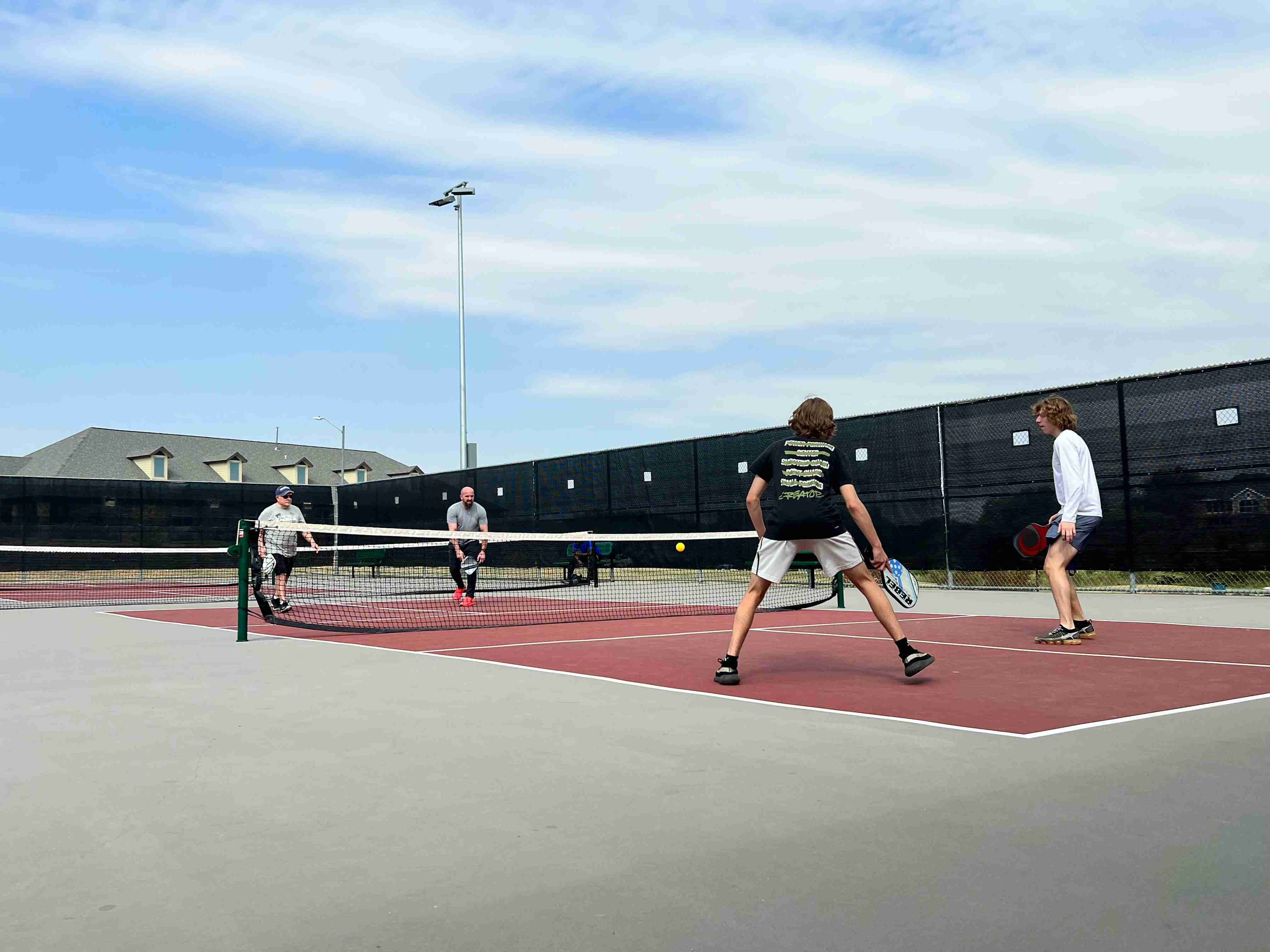 Four players engaged in a pickleball game on an outdoor court, demonstrating the double bounce rule. Two teams of two are positioned on each side of the net, actively playing. The court is marked for pickleball with players in motion, emphasizing the rule where the ball must bounce once on each side before volleys can be played.