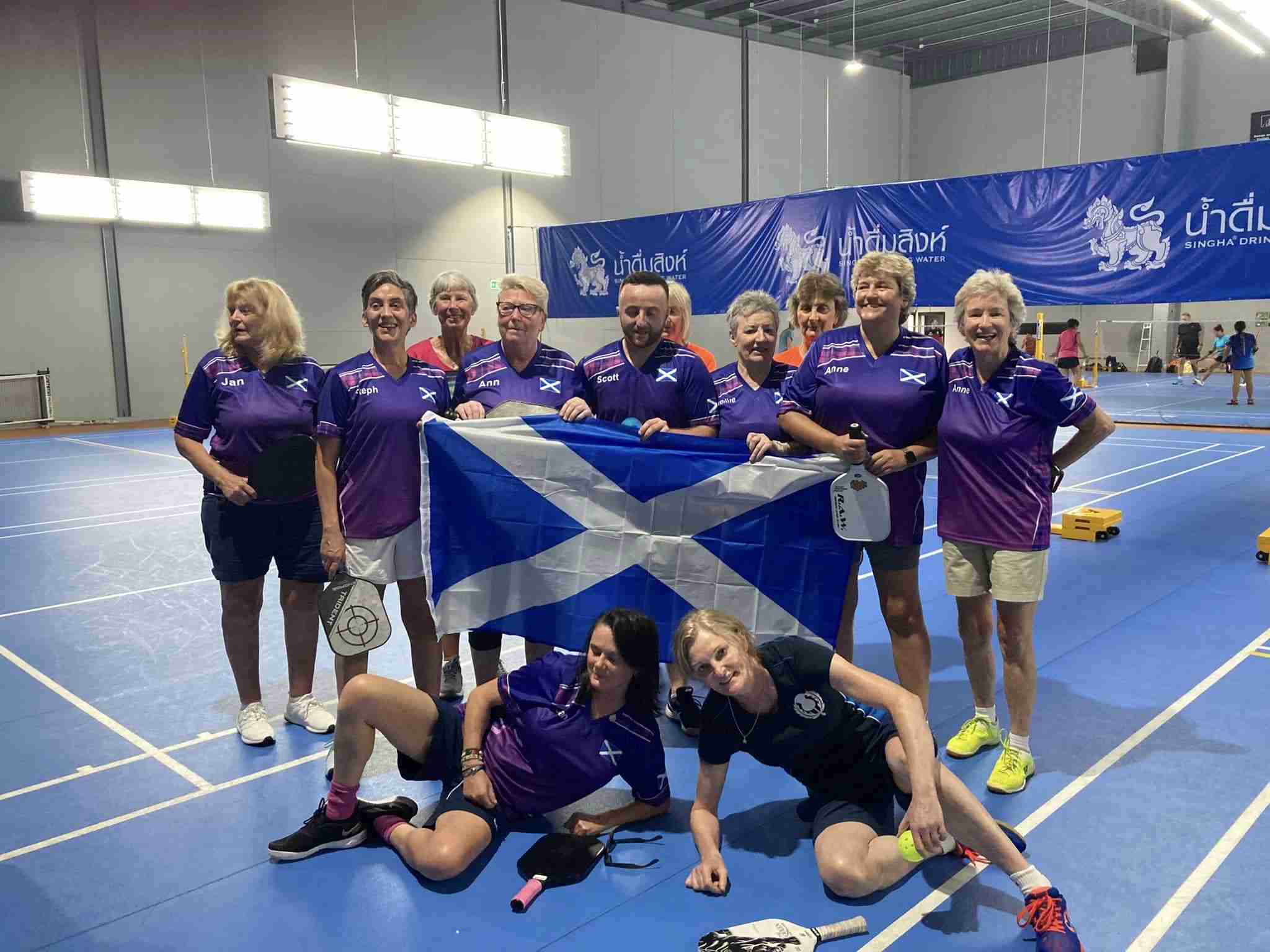 A group of pickleball players wearing matching purple uniforms, holding a Scottish flag, poses on an indoor court.
