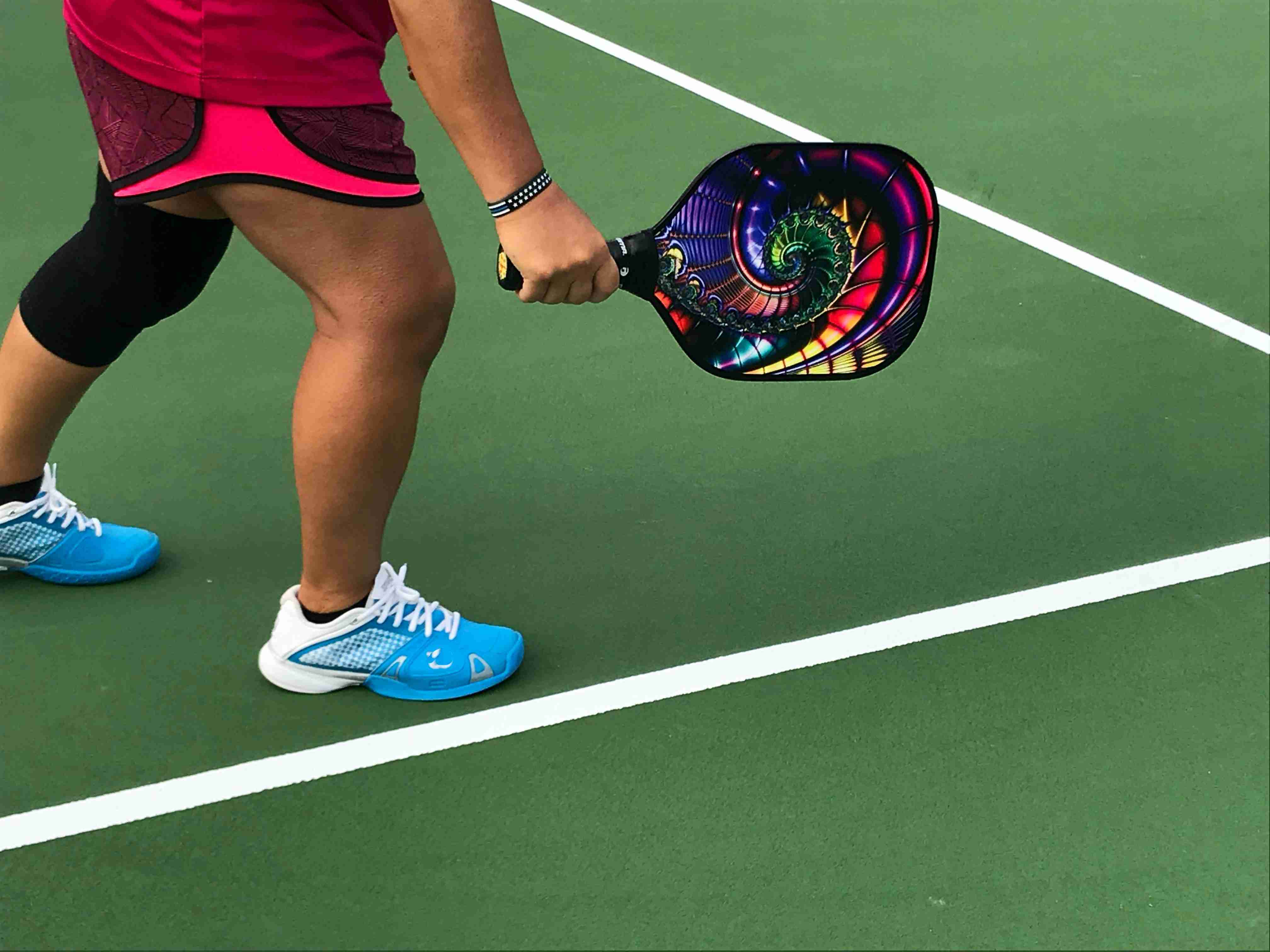 Close-up of a pickleball player holding a colorful paddle and wearing pickleball shoes while standing on a green court.
