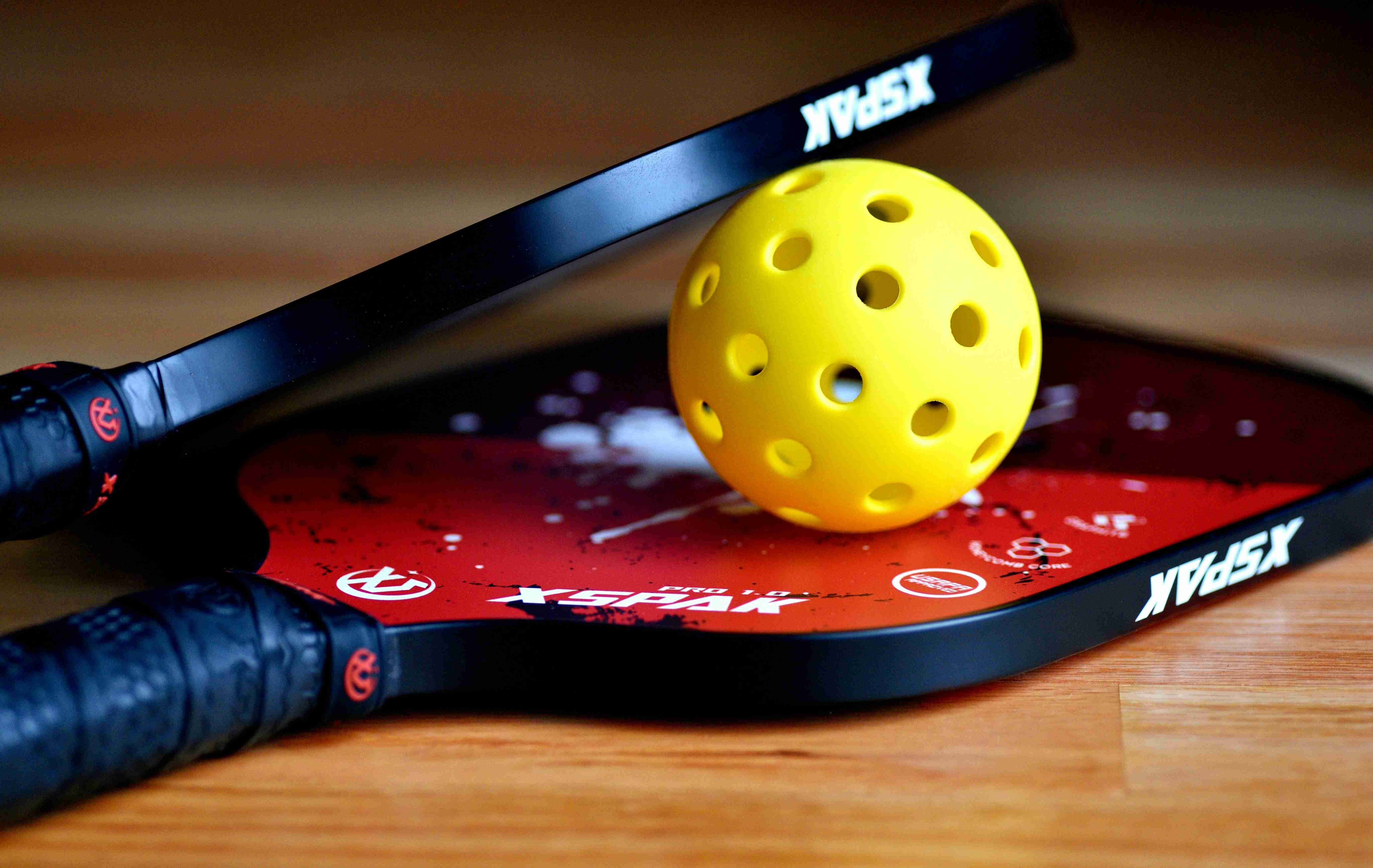 Pickleball paddle and yellow pickleball ball on a wooden floor, featuring the XSPAK paddle brand