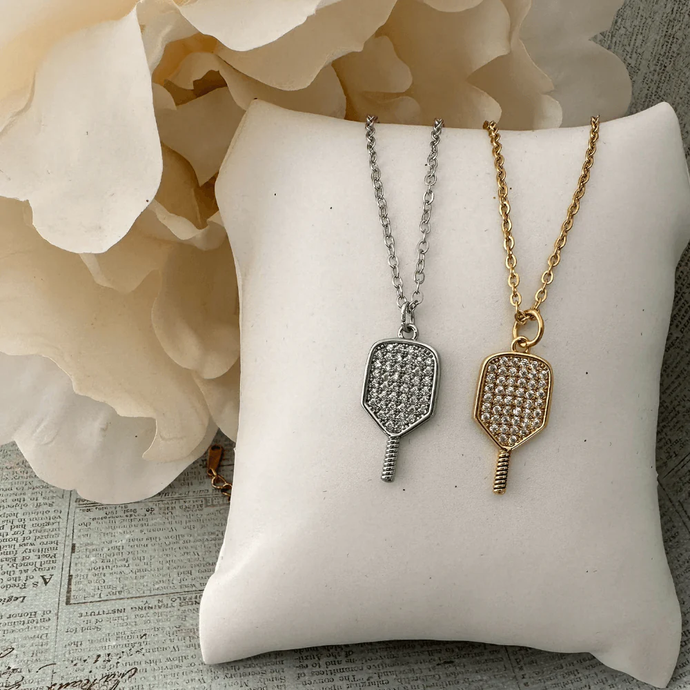 Elegant pickleball jewelry featuring silver and gold pickleball paddle necklaces adorned with sparkling crystals, displayed on a soft cushion background.