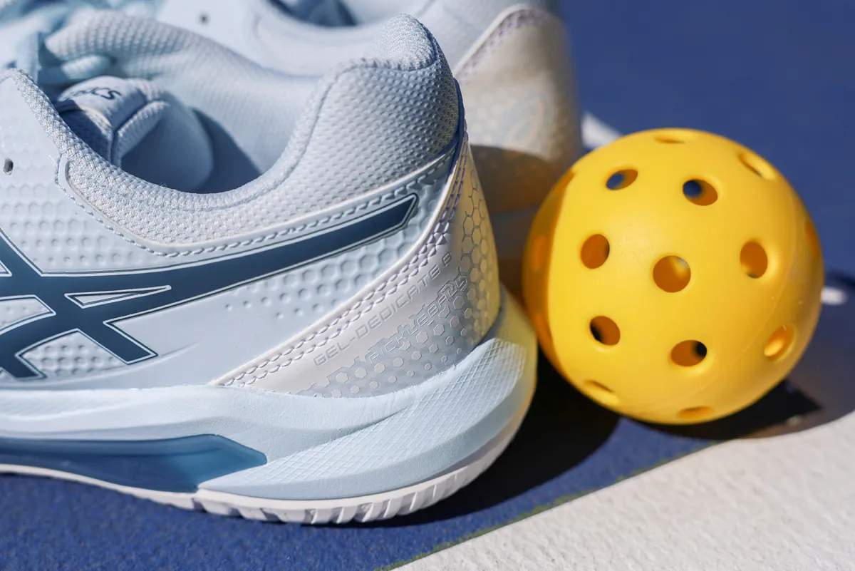 Close-up of a light blue pickleball shoe next to a yellow pickleball with holes, on a blue court surface.