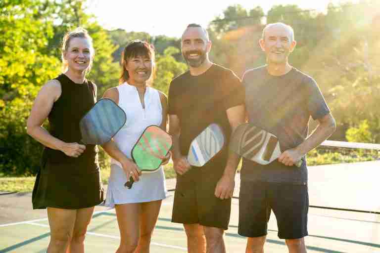 Four smiling pickleball players holding colorful pickleball paddles on an outdoor court, standing together in warm sunlight, ready for a game.