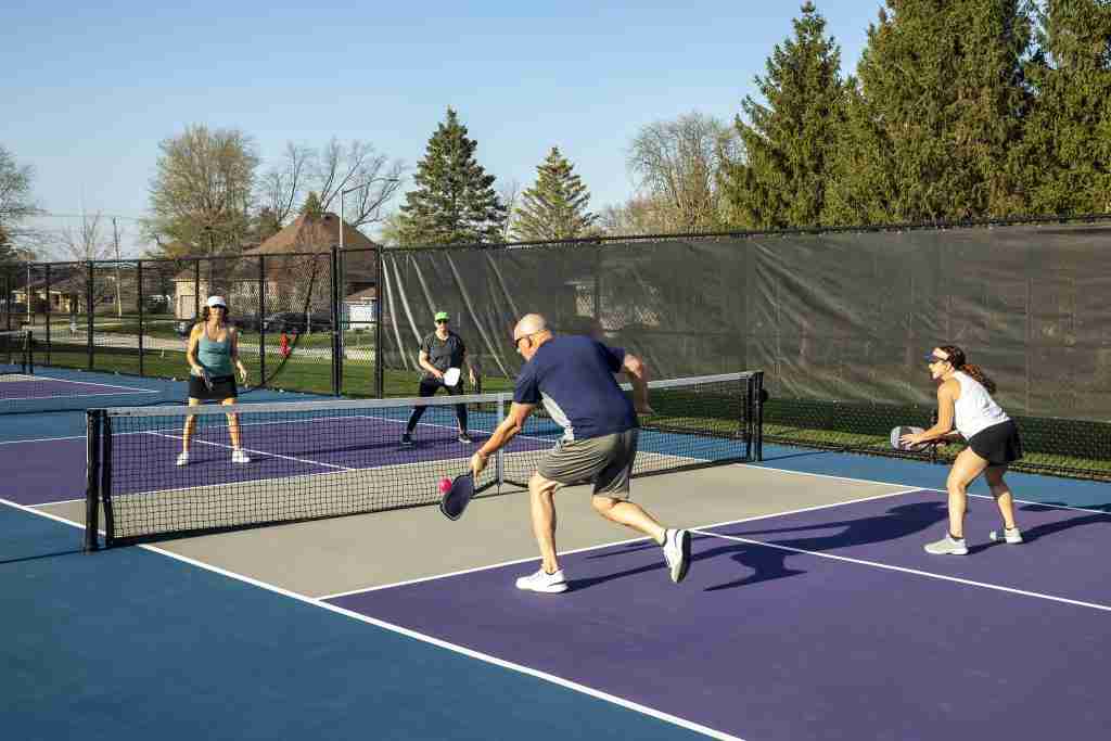 Four players engaged in a doubles pickleball game on a sunny day, showcasing active play and strategic positioning while adhering to the double-bounce rule.