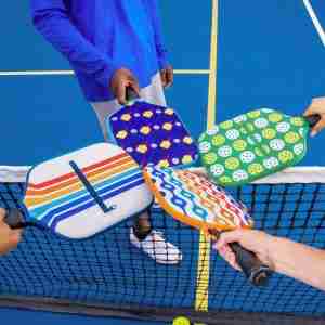 Four players stand around a pickleball net, each holding a pickleball paddle with a colorful pickleball paddle cover featuring different patterns. The covers include designs with stripes, pickleball paddles and balls, and abstract waves. The players' hands and paddles meet in the center of the image, symbolizing the start of a game on a blue pickleball court.