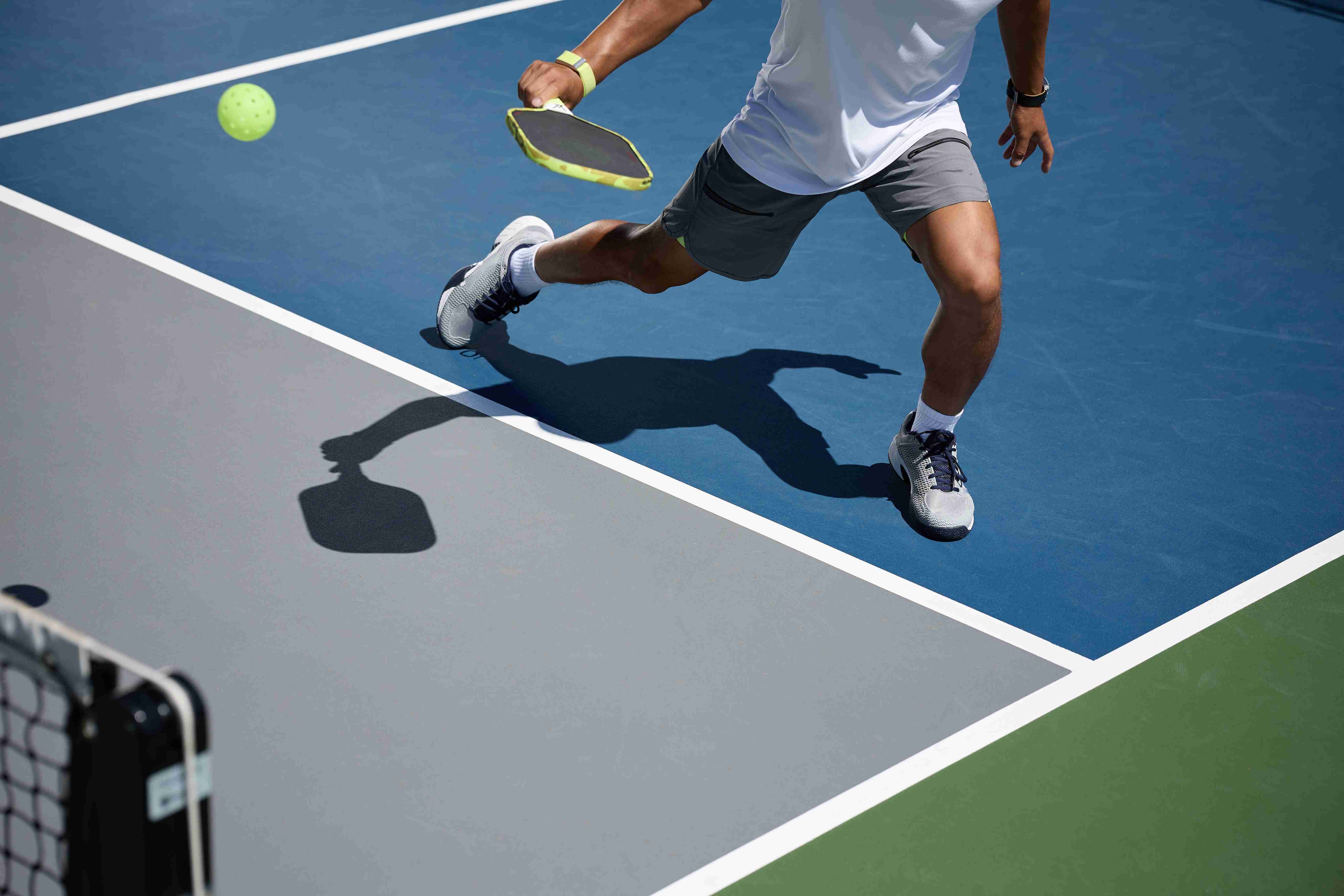 Player demonstrating strong pickleball footwork while lunging for a shot near the non-volley zone on a blue and grey court.