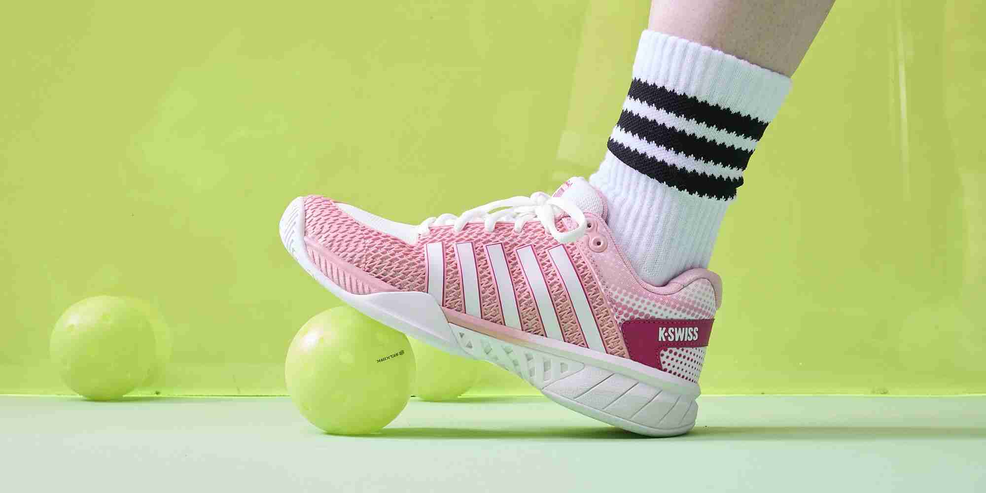 K-Swiss pickleball shoes in pink worn by a player balancing on a pickleball court with neon balls in the background