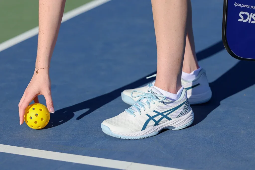 Woman wearing ASICS outdoor pickleball shoes picking up a yellow pickleball on an outdoor court, showcasing grip and stability.