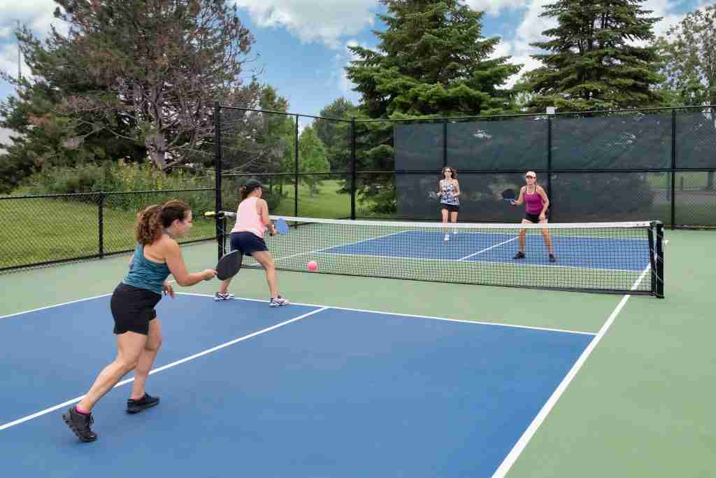Four women playing doubles on an outdoor court, demonstrating the rules of pickleball doubles with teamwork, positioning, and strategy near the net.