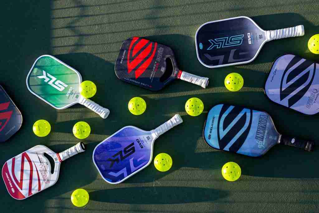 An image of multiple Selkirk paddles in various models (including EVO, Invikta, VANGUARD, and Project Boomstik) and colors, along with several neon yellow pickleballs, all arranged on a green pickleball court surface. The paddles showcase different shapes—from elongated to traditional widebody—highlighting the diversity of Selkirk paddles for all play styles.